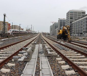 Rechts und links Gebäude, in der Mitte vier Gleise. Rechts ein ICE, auf einem Gleis ein Bagger.