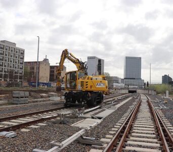 Ein Bagger auf einem Gleis vor einem Tunnelprotal, im Hintergrund Gebäude